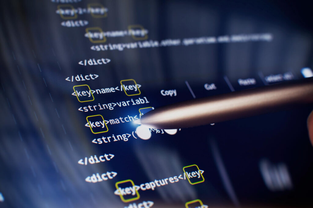 Close-up of a computer screen displaying code with highlighted syntax, while a hand holds a stylus, pointing towards the code—suggesting programming, software development, or analyzing threats like ransomware. | BeMyNet.com