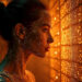 A woman with glowing dots on her skin gazes intently at an illuminated grid of orange lights, embodying the strength of a Human Firewall and creating a futuristic, ethereal atmosphere. | BeMyNet.com